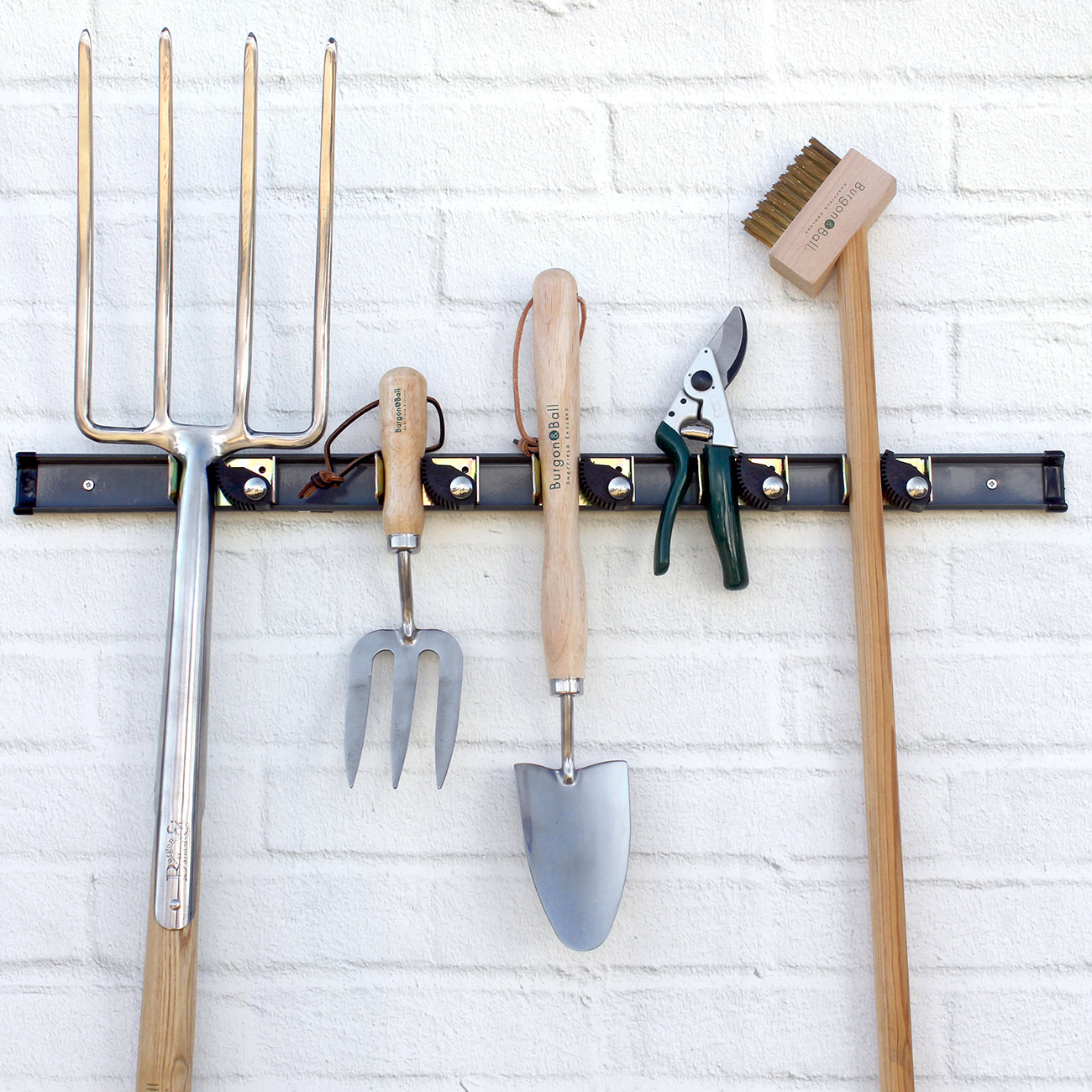 Set of gardening tools on a wall-mounted rack against a white brick wall.
