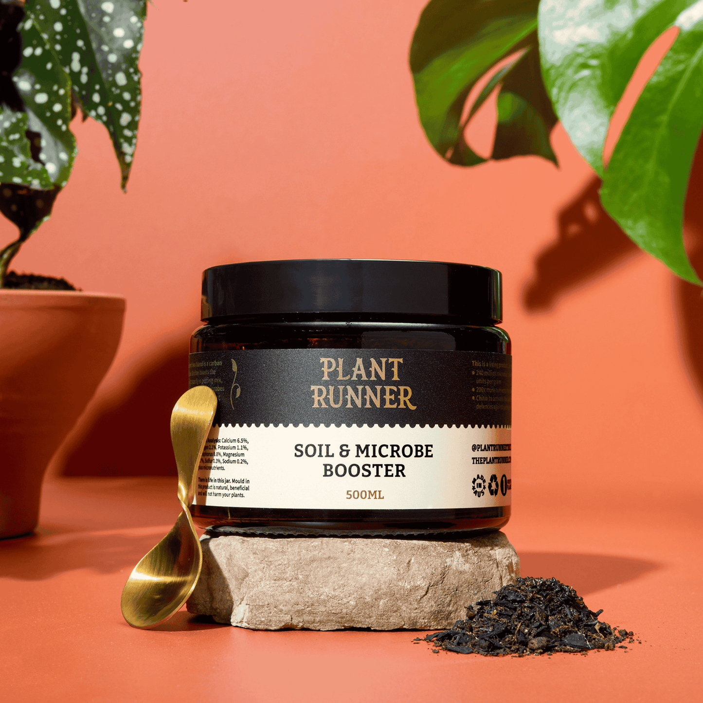 Jar of 'Plant Runner Soil & Microbe Booster' on a stone block with plants in the background.