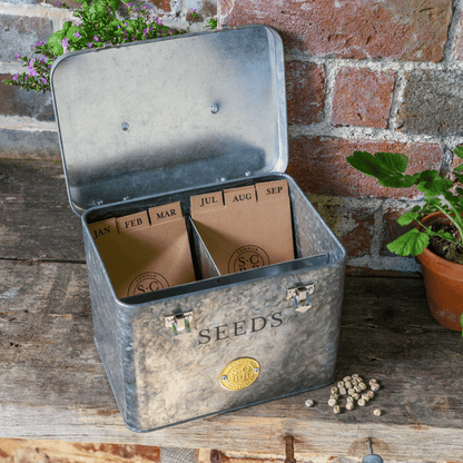 Metal seed storage box with labeled compartments on a wooden surface against a brick wall.
