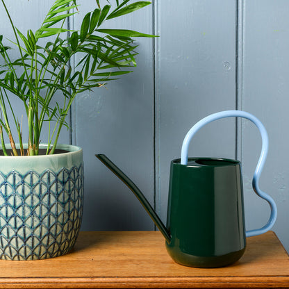 Dark green Burgon & Ball indoor watering can next to a potted plant on a wooden surface.