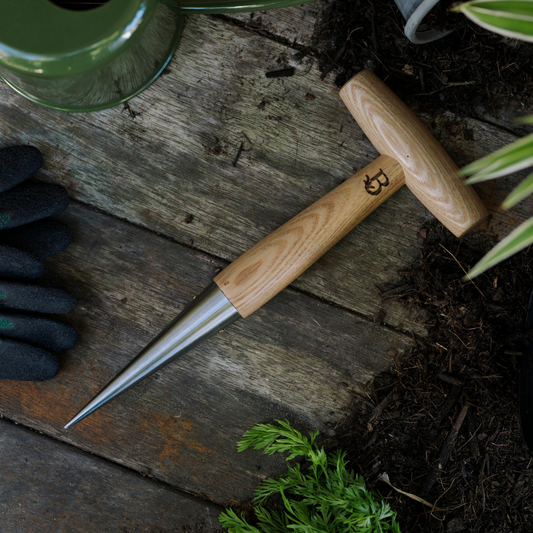 Gardening dibber with wooden handle on a wooden surface with soil and plants.