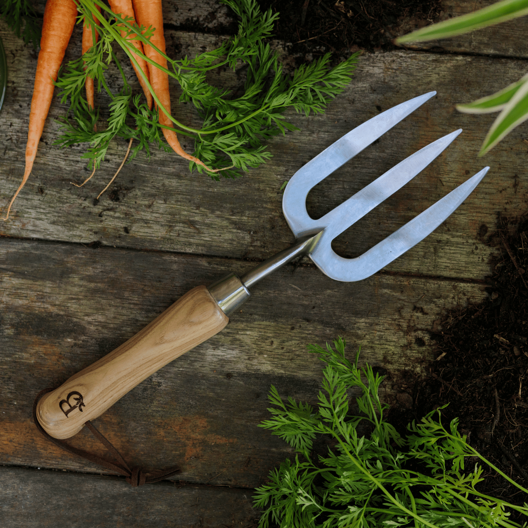 'Get Growing' gardening fork with wooden handle on a wooden surface with carrots and greenery.