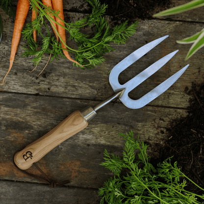 'Get Growing' gardening fork with wooden handle on a wooden surface with carrots and greenery.